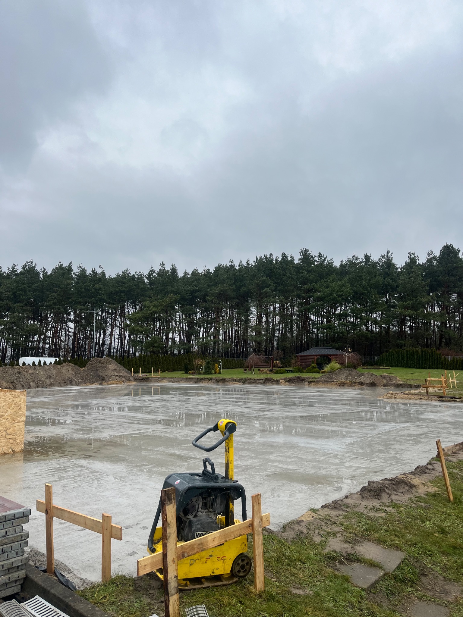 Świeżo wylana płyta betonowa pod budowę domu, widoczna zagęszczarka wibracyjna, drewniane szalunki, w tle las i pochmurne niebo.