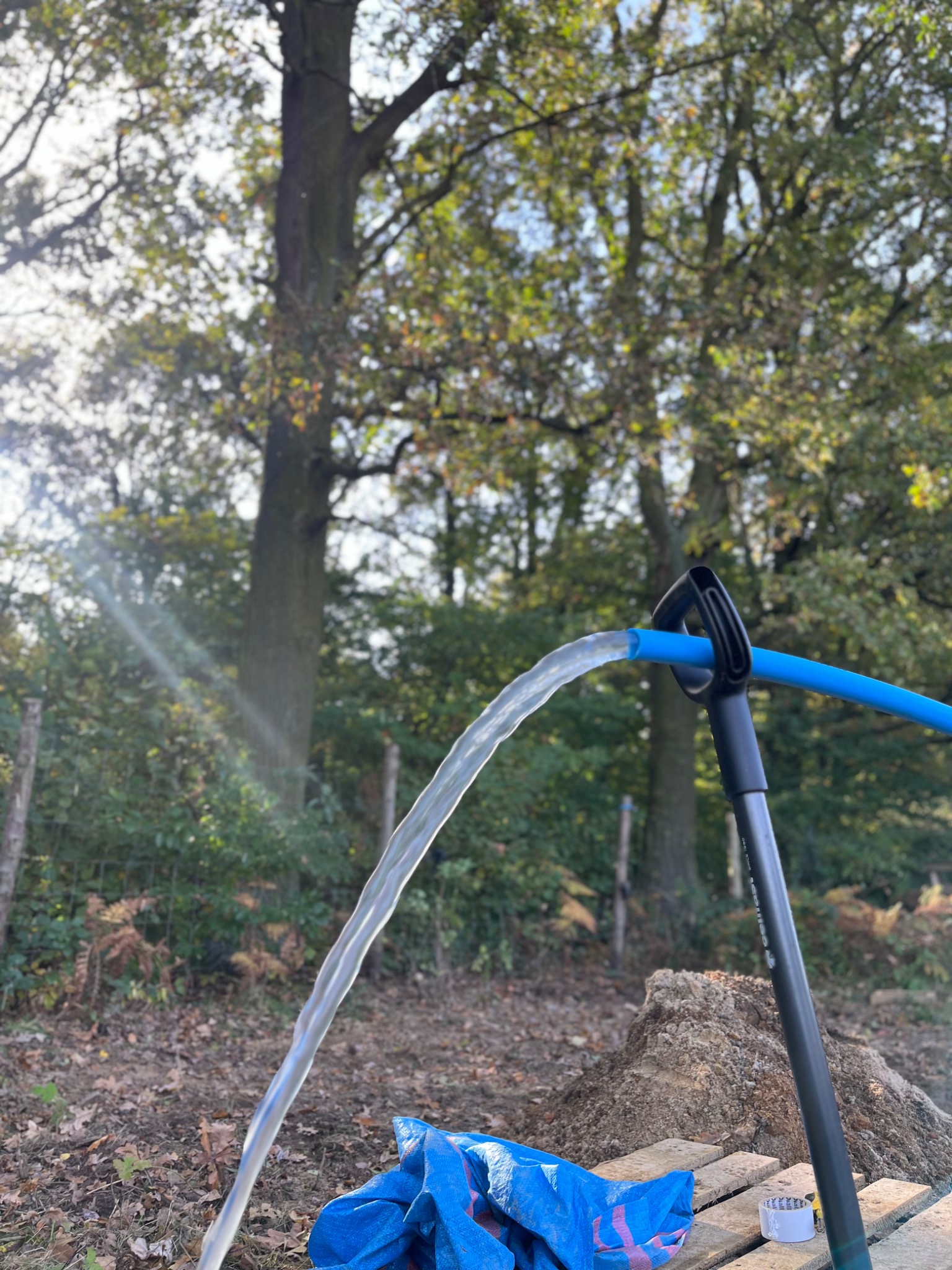 Strumień wody wypływa z niebieskiego węża zamontowanego na czarnym stojaku, w tle ziemia, drzewa i niebieska plandeka.