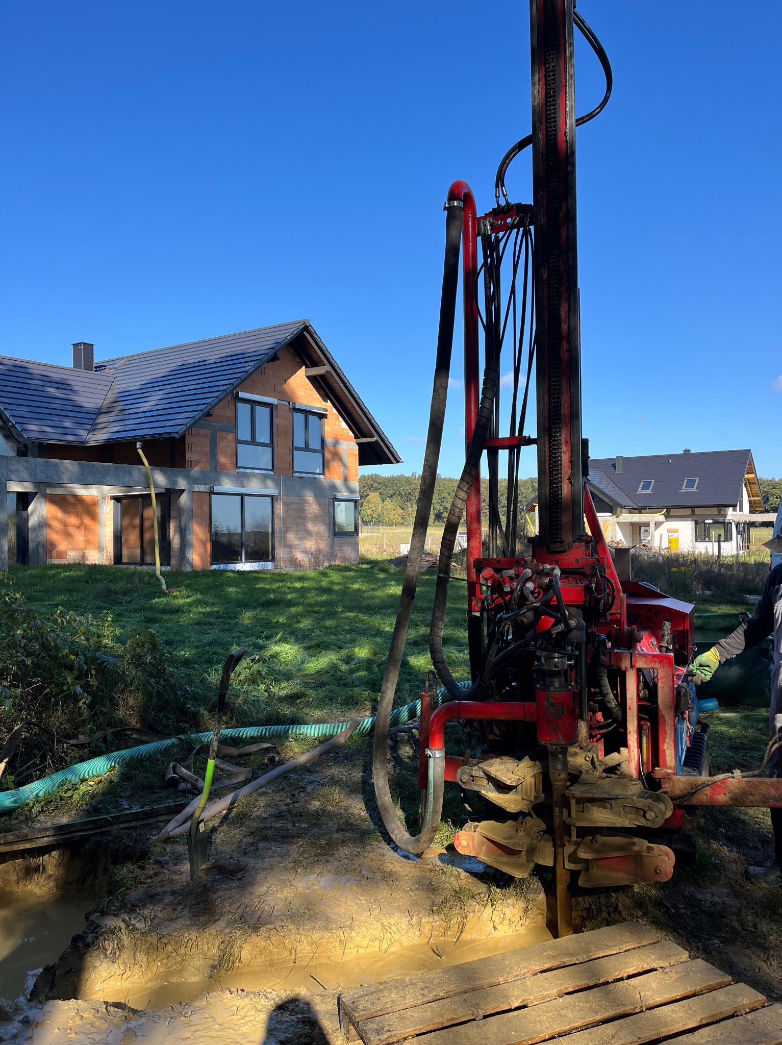 Czerwona wiertnica do studni głębinowych na tle budowy domu jednorodzinnego i błękitnego nieba, z widocznym wykopem wypełnionym wodą.