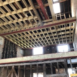 LewBud - Wnętrze budynku w trakcie remontu, z widoczną konstrukcją stropu z drewna i metalu, ściany z odsłoniętą starą cegłą i fragmentami tynku, oraz widocznym oknem w tle.