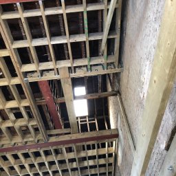 LewBud - Konstrukcja drewniana stropu z elementami stalowymi w trakcie montażu, widoczne fragmenty starej ceglanej ściany i okno w dolnej części kadru.