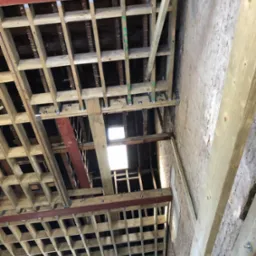 Konstrukcja drewniana stropu z elementami stalowymi w trakcie montażu, widoczne fragmenty starej ceglanej ściany i okno w dolnej części kadru.