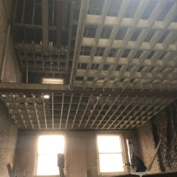 LewBud - Wnętrze w trakcie remontu: widoczne drewniane elementy konstrukcyjne sufitu, gołe ceglane ściany, okna z intensywnym światłem dziennym, drabina i narzędzia budowlane.