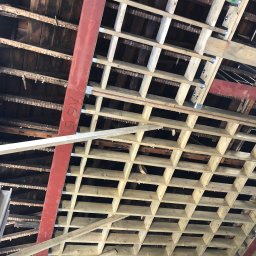 LewBud - Sufit w trakcie budowy z widoczną konstrukcją drewnianą, wzmocniony czerwoną, stalową belką z napisem 'A5 51', stare drewniane deski widoczne pod spodem.