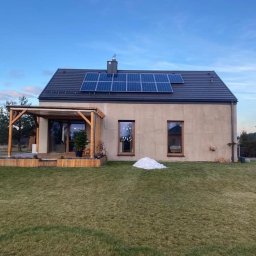 LewBud - Nowoczesny dom z beżową elewacją, panelami słonecznymi na dachu i drewnianą pergolą z tarasem, otoczony trawnikiem i skąpany w popołudniowym świetle.