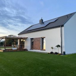 LewBud - Nowoczesny dom jednorodzinny z białymi ścianami, panelami słonecznymi na dachu i drewnianą pergolą z huśtawką oraz fragmentami elewacji wykończonymi cegłą klinkierową.