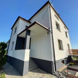 LewBud - Dwukondygnacyjny dom po renowacji elewacji w kolorach bieli i grafitu, z jasną kostką brukową i elementami zieleni w słoneczny dzień.