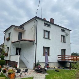 LewBud - Dwupiętrowy dom z białymi ścianami, brązowymi ramami okiennymi i drewnianym balkonem, widziany z perspektywy ogrodu z kostką brukową i parasolem przeciwsłonecznym.