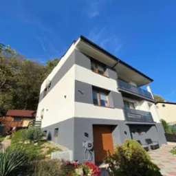 Nowoczesny dom z elewacją w szaro-białe pasy, z balkonami z metalowymi barierkami, otoczony zielenią i kostką brukową, widok z dołu na tle błękitnego nieba.