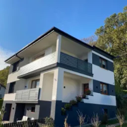 Nowoczesny, trzypiętrowy dom z elewacją w kolorach bieli i grafitu, z drewnianymi oknami, balkonami z poziomymi balustradami, kwiatami w donicach na schodach wejściowych i niskim, czarnym ogrodzeniem.