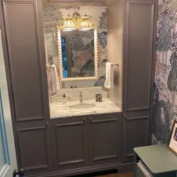 Elegancka łazienka z zabudową szafkową w kolorze taupe, marmurowym blatem umywalkowym, lustrem w ramie z pereł i tapetą w motywy leśne, oświetlona lampami z plisowanymi abażurami.