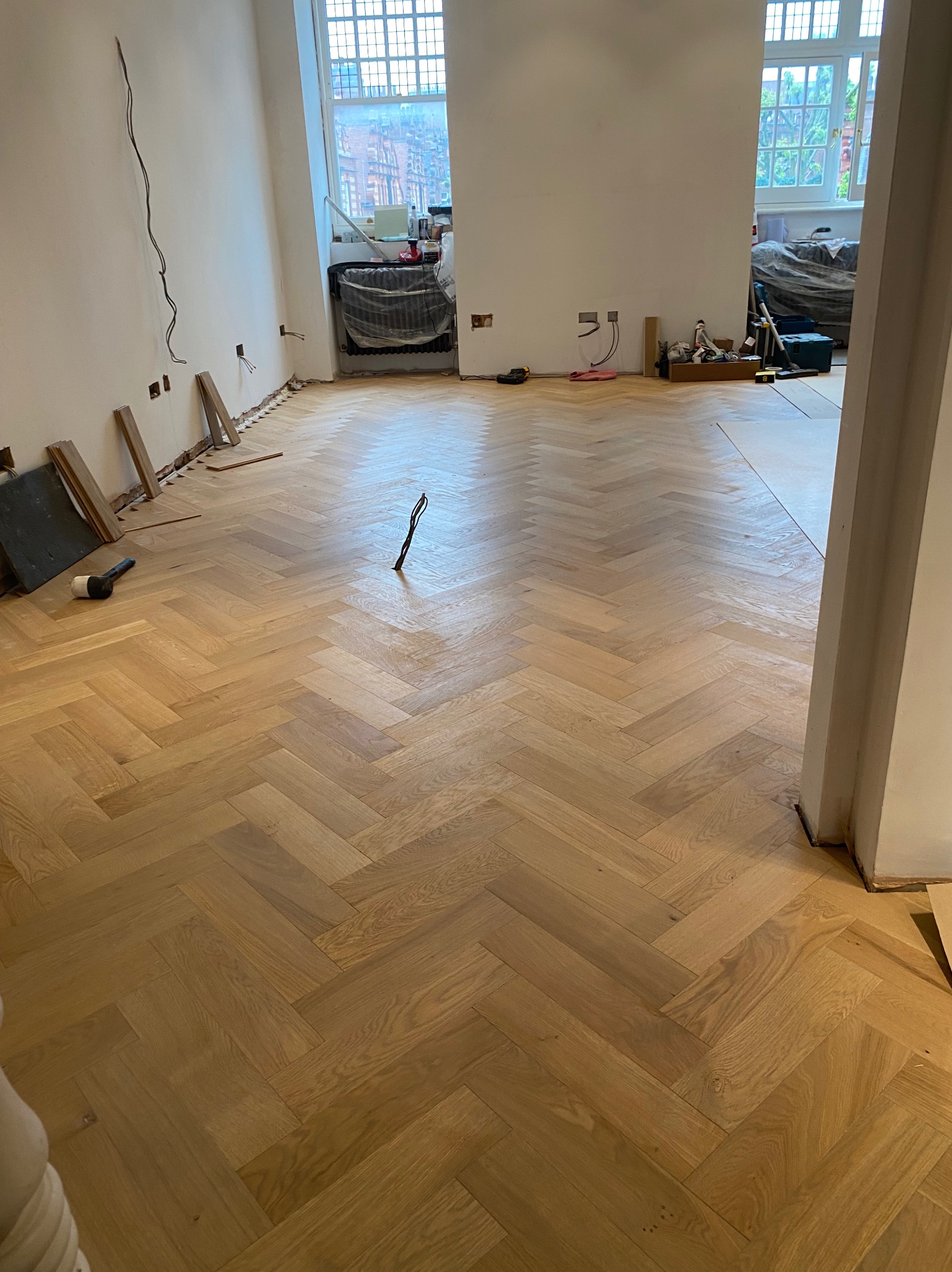 Układanie parkietu w jodełkę w jasnym odcieniu drewna, widoczne niedokończone ściany, wystające przewody elektryczne, gumowy młotek i fragmenty desek pod ścianą.