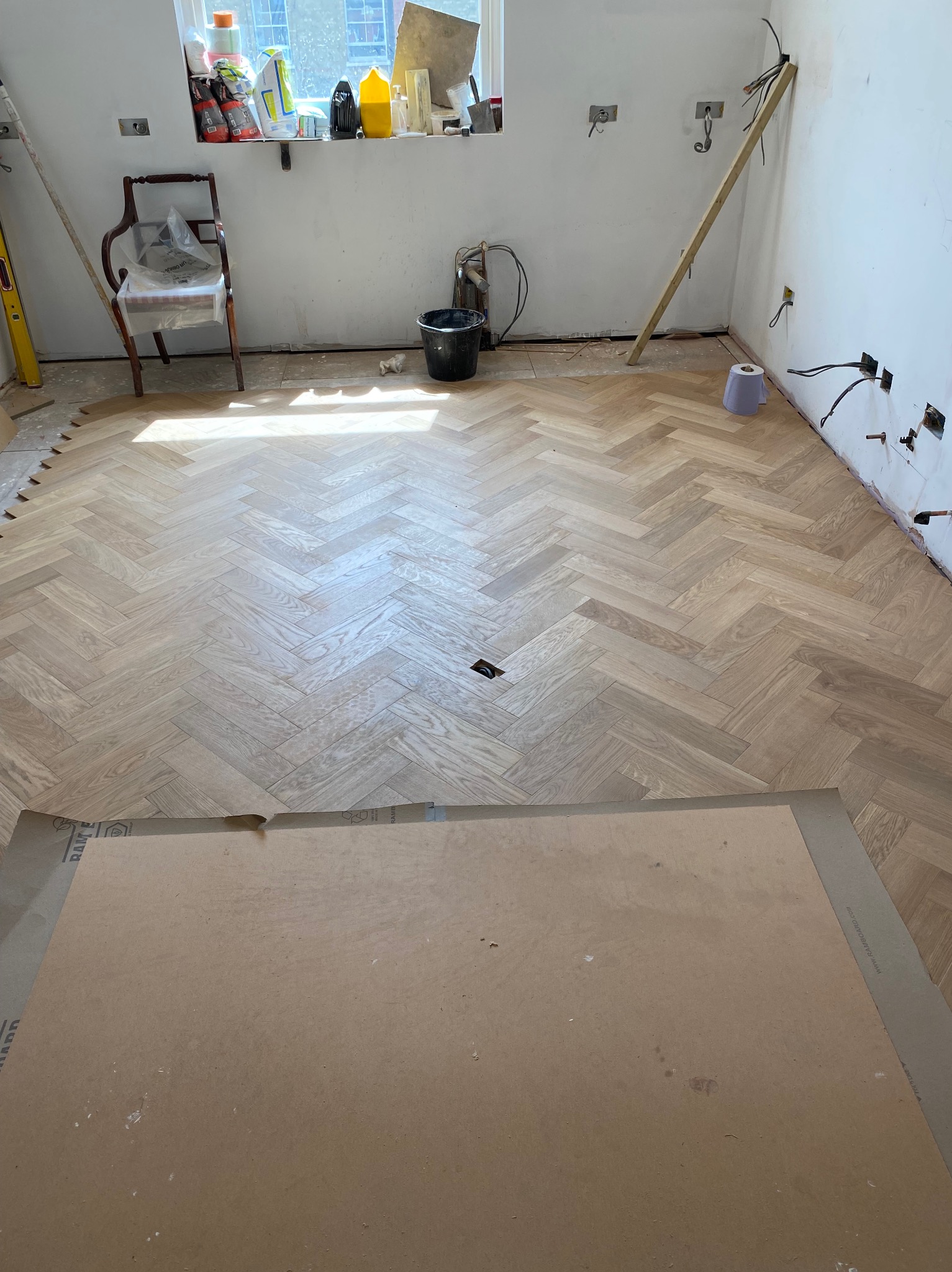 Jasna, drewniana podłoga ułożona w jodełkę w trakcie montażu w pomieszczeniu z widocznymi instalacjami elektrycznymi i przygotowanymi ścianami, częściowo zakryta kartonem ochronnym.