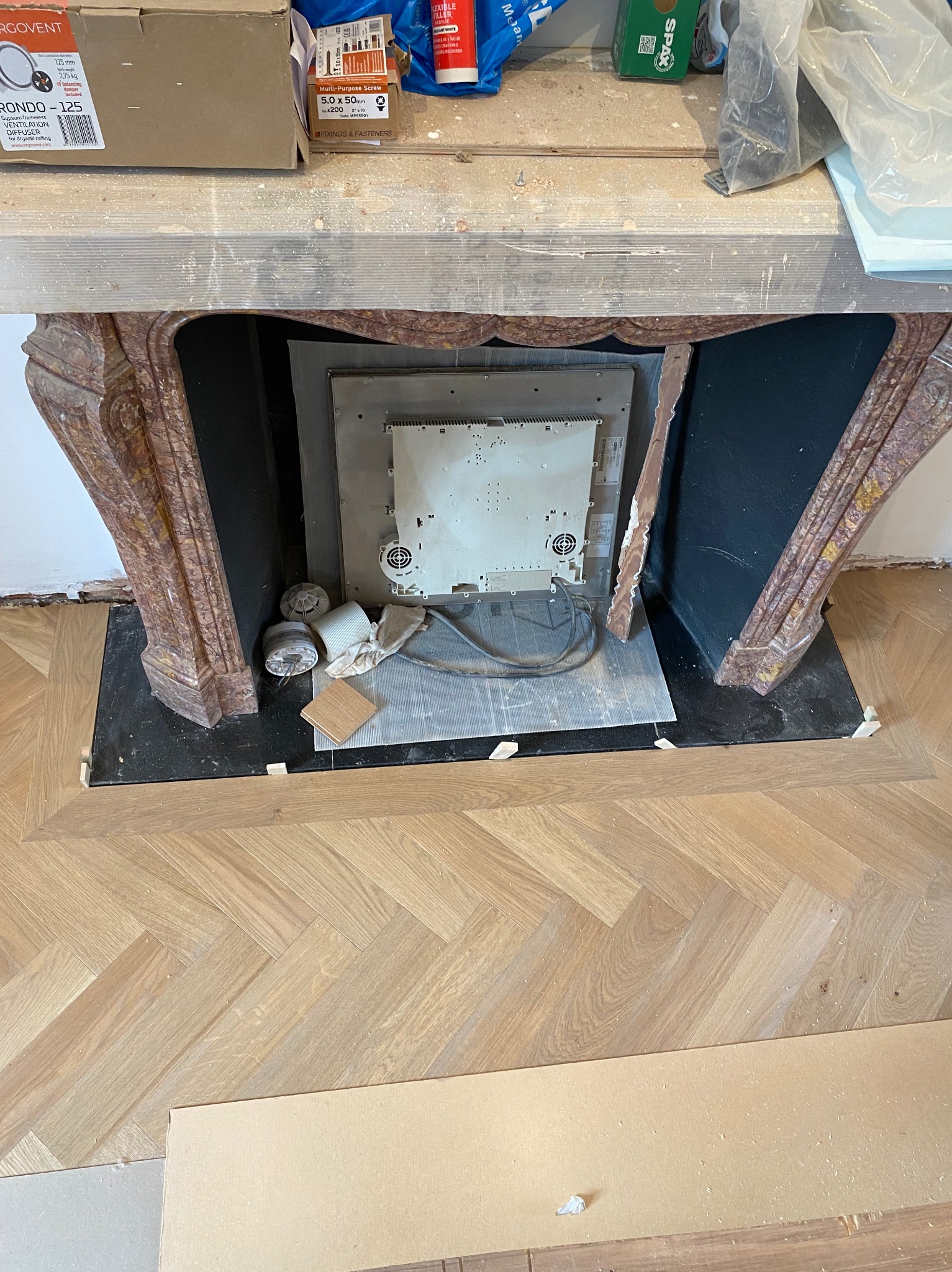 Fragment demontażu lub montażu elektrycznego wkładu kominkowego w marmurowej obudowie, widoczne kable, podłączenia i elementy mocujące, na podłodze parkiet w jodełkę, w tle kartony i materiały...