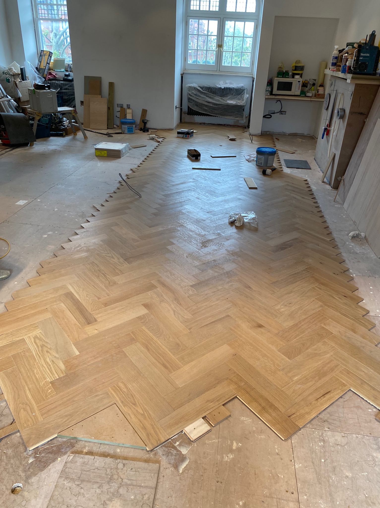 Układanie parkietu w jodełkę w przestronnym pokoju; widoczne deski parkietu ułożone na znacznym obszarze, a także nieułożone elementy i narzędzia pracy, wskazujące na trwający proces montażu.