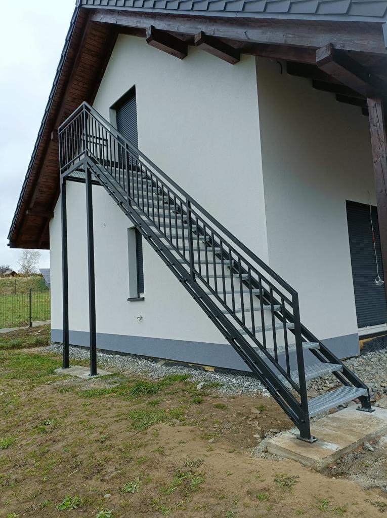 Zewnętrzne stalowe schody z ażurowymi stopniami prowadzące do drzwi na piętrze, biała elewacja budynku z szarą podmurówką, widoczne okno z żaluzjami.