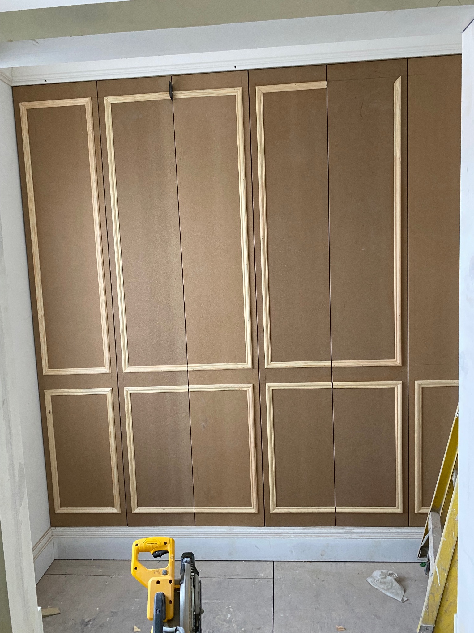 Montaż dekoracyjnych paneli ściennych z MDF z drewnianymi listwami; żółta piła ukośnica na podłodze, drabina w tle.