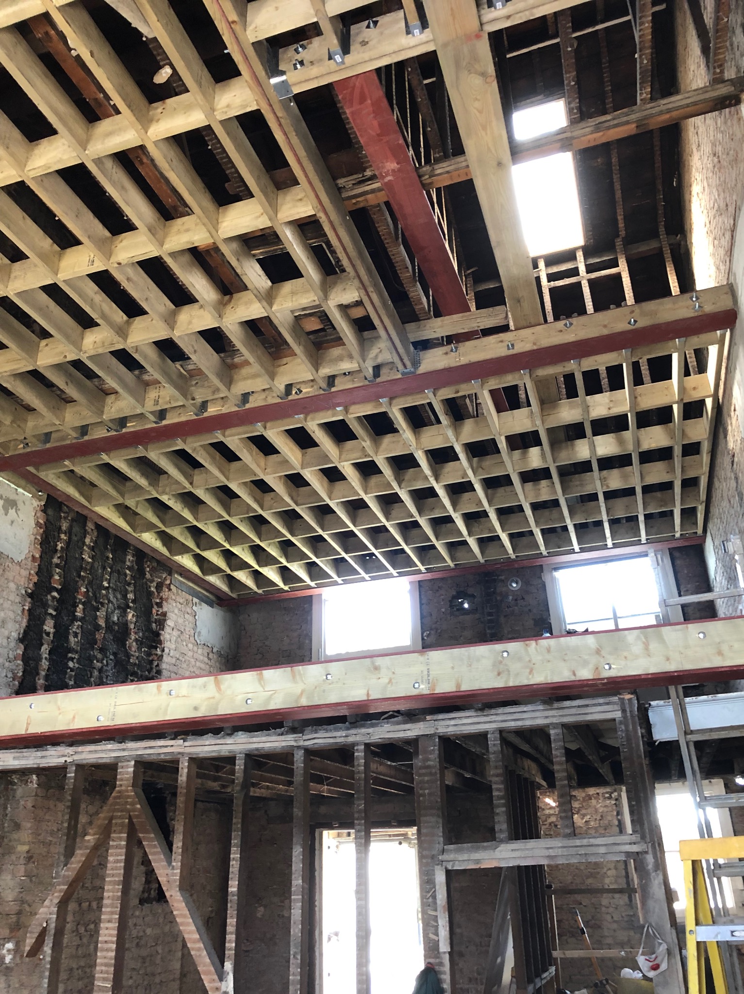 Wnętrze budynku w trakcie remontu, z widoczną konstrukcją stropu z drewna i metalu, ściany z odsłoniętą starą cegłą i fragmentami tynku, oraz widocznym oknem w tle.