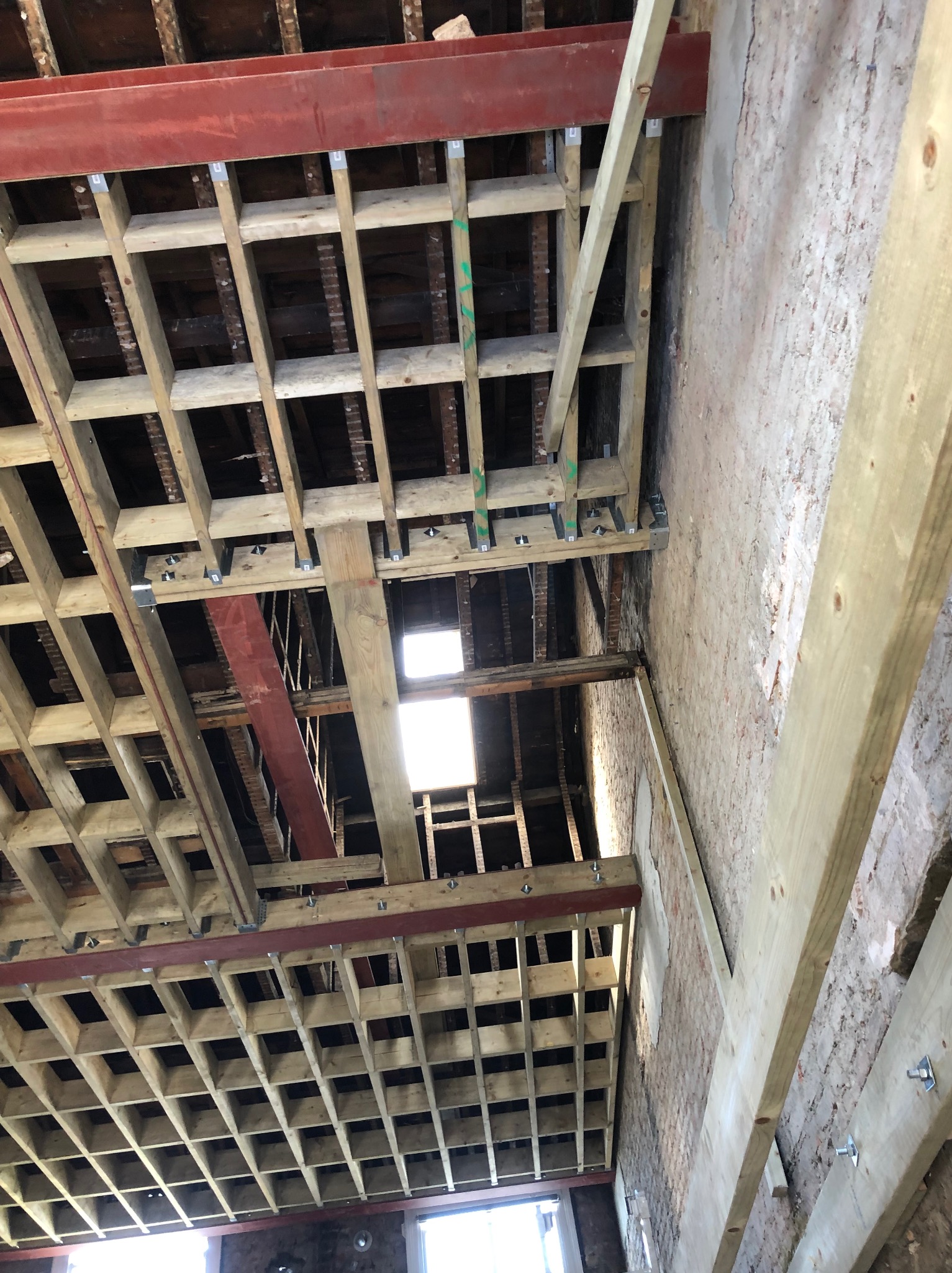 Konstrukcja drewniana stropu z elementami stalowymi w trakcie montażu, widoczne fragmenty starej ceglanej ściany i okno w dolnej części kadru.