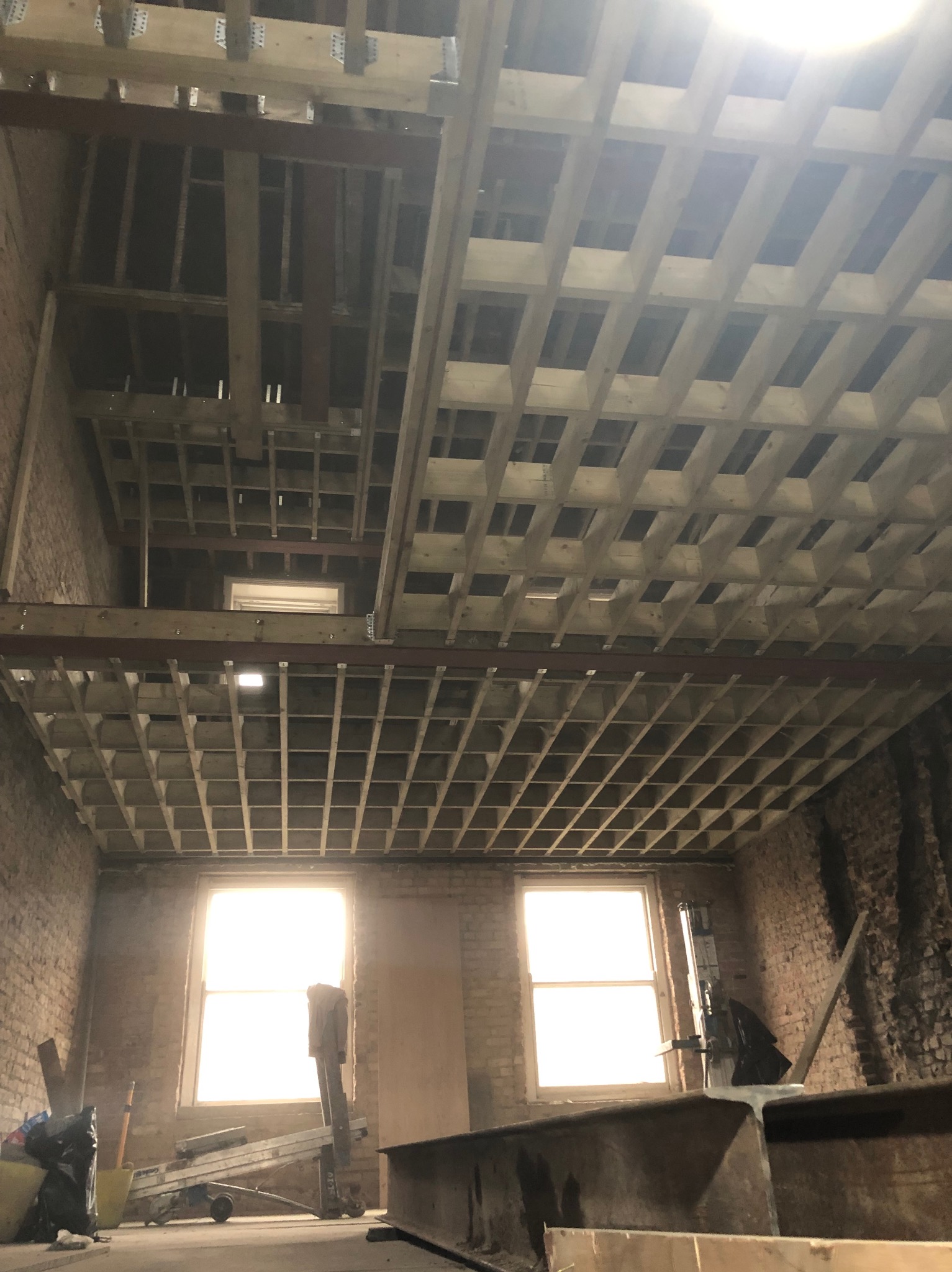 Wnętrze w trakcie remontu: widoczne drewniane elementy konstrukcyjne sufitu, gołe ceglane ściany, okna z intensywnym światłem dziennym, drabina i narzędzia budowlane.