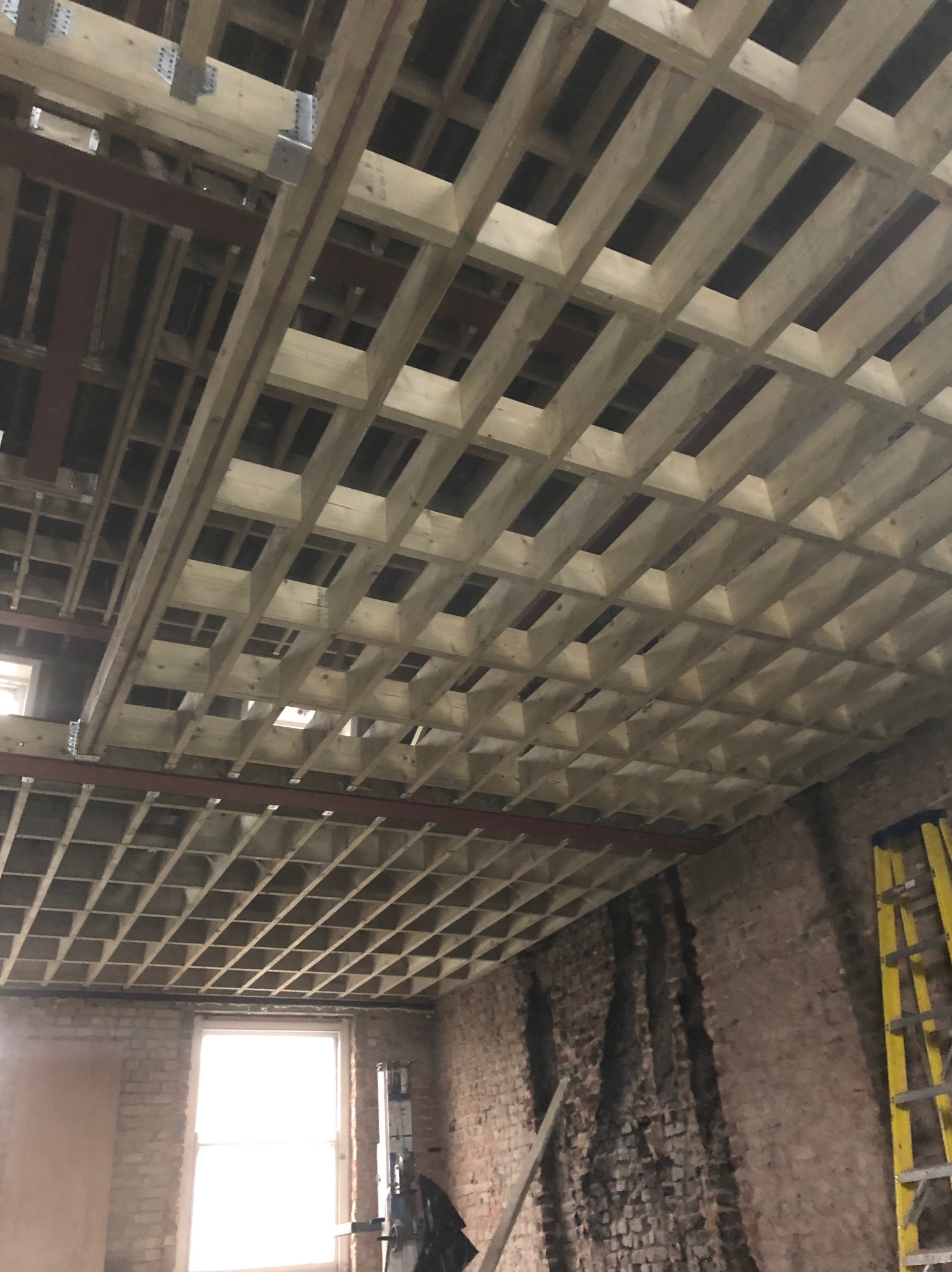 Drewniana konstrukcja sufitu podwieszanego z elementami metalowymi, widok z dołu, surowe ściany z cegły w tle, żółta drabina oparta o ścianę.