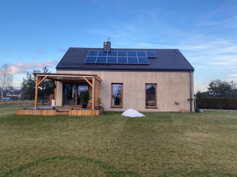 Nowoczesny dom z beżową elewacją, panelami słonecznymi na dachu i drewnianą pergolą z tarasem, otoczony trawnikiem i skąpany w popołudniowym świetle.