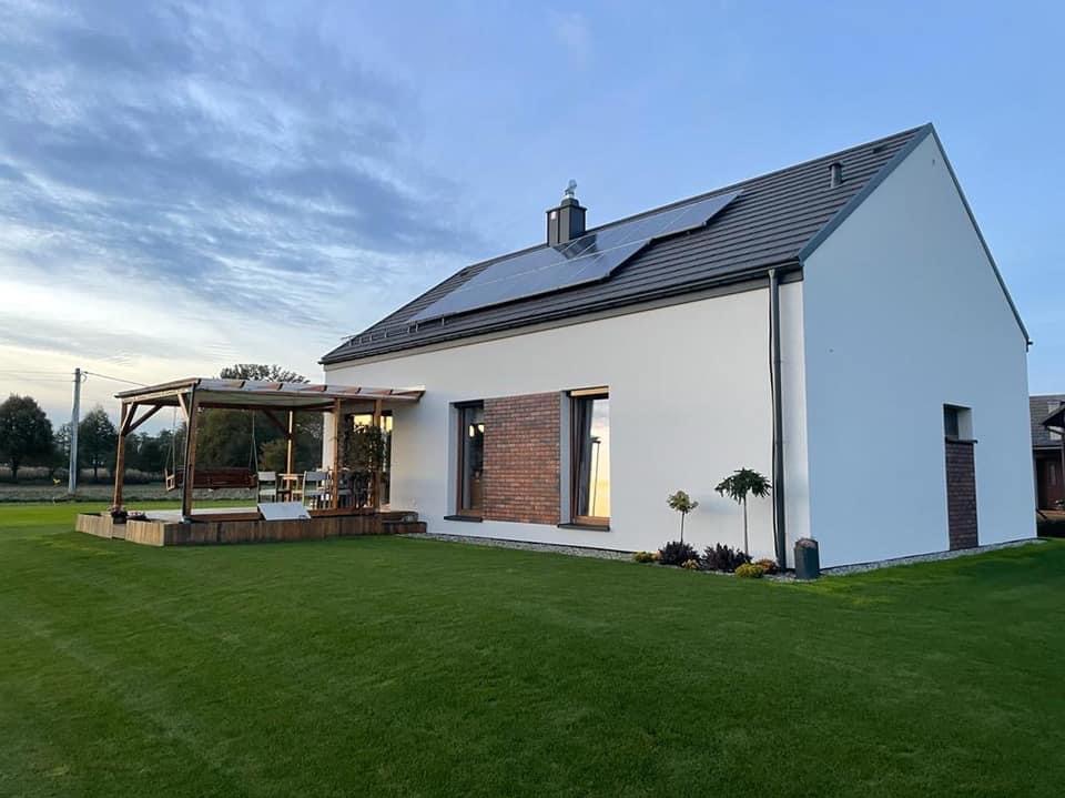 Nowoczesny dom jednorodzinny z białymi ścianami, panelami słonecznymi na dachu i drewnianą pergolą z huśtawką oraz fragmentami elewacji wykończonymi cegłą klinkierową.