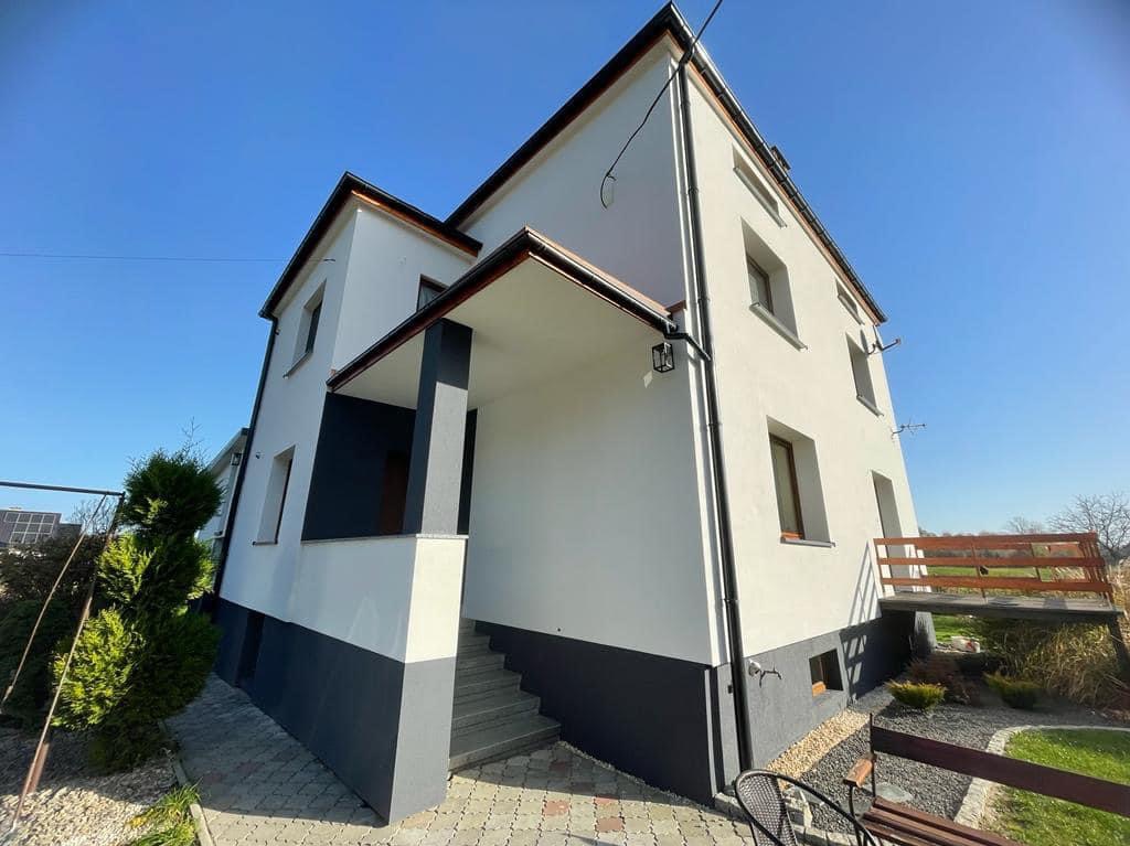 Dwukondygnacyjny dom po renowacji elewacji w kolorach bieli i grafitu, z jasną kostką brukową i elementami zieleni w słoneczny dzień.