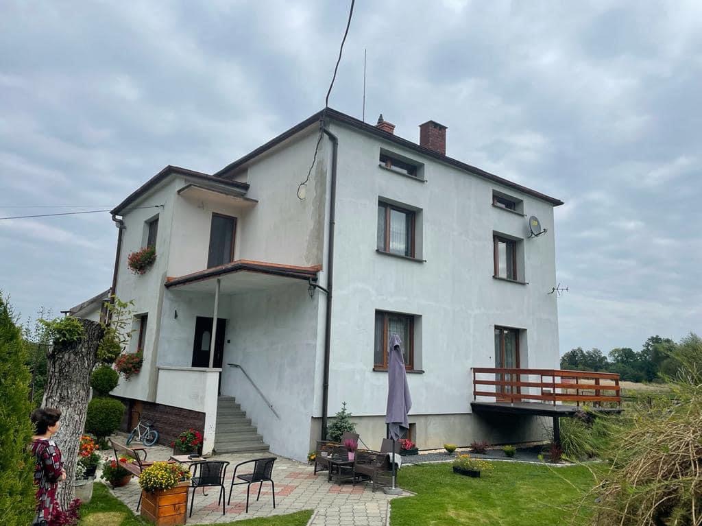 Dwupiętrowy dom z białymi ścianami, brązowymi ramami okiennymi i drewnianym balkonem, widziany z perspektywy ogrodu z kostką brukową i parasolem przeciwsłonecznym.