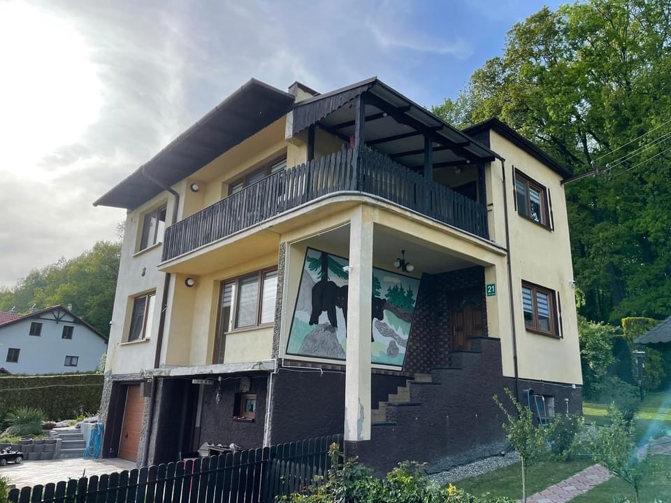 Dwupiętrowy dom z balkonem i muralem przedstawiającym niedźwiedzie na elewacji, widok z dołu na tle zieleni.