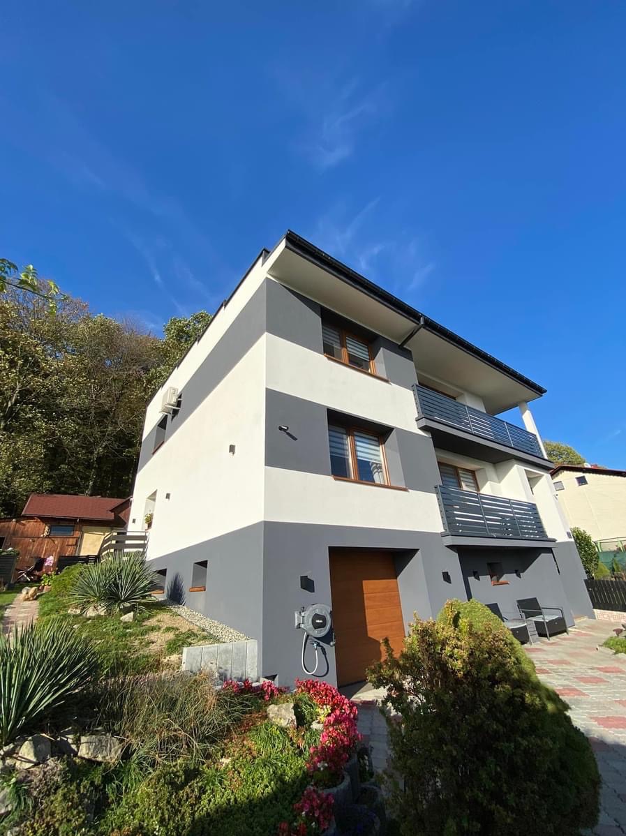 Nowoczesny dom z elewacją w szaro-białe pasy, z balkonami z metalowymi barierkami, otoczony zielenią i kostką brukową, widok z dołu na tle błękitnego nieba.