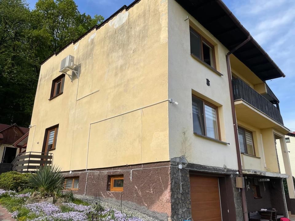 Dwukondygnacyjny dom z elewacją w odcieniach beżu i brązu, z widocznym klimatyzatorem na ścianie i fragmentem balkonu, otoczony zielenią i fioletowymi kwiatami.