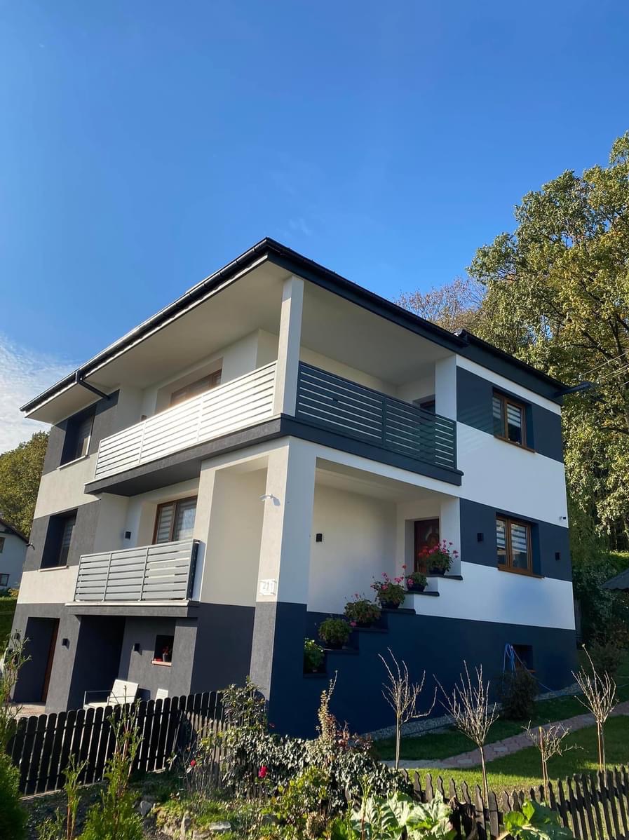 Nowoczesny, trzypiętrowy dom z elewacją w kolorach bieli i grafitu, z drewnianymi oknami, balkonami z poziomymi balustradami, kwiatami w donicach na schodach wejściowych i niskim, czarnym ogrodzeniem.