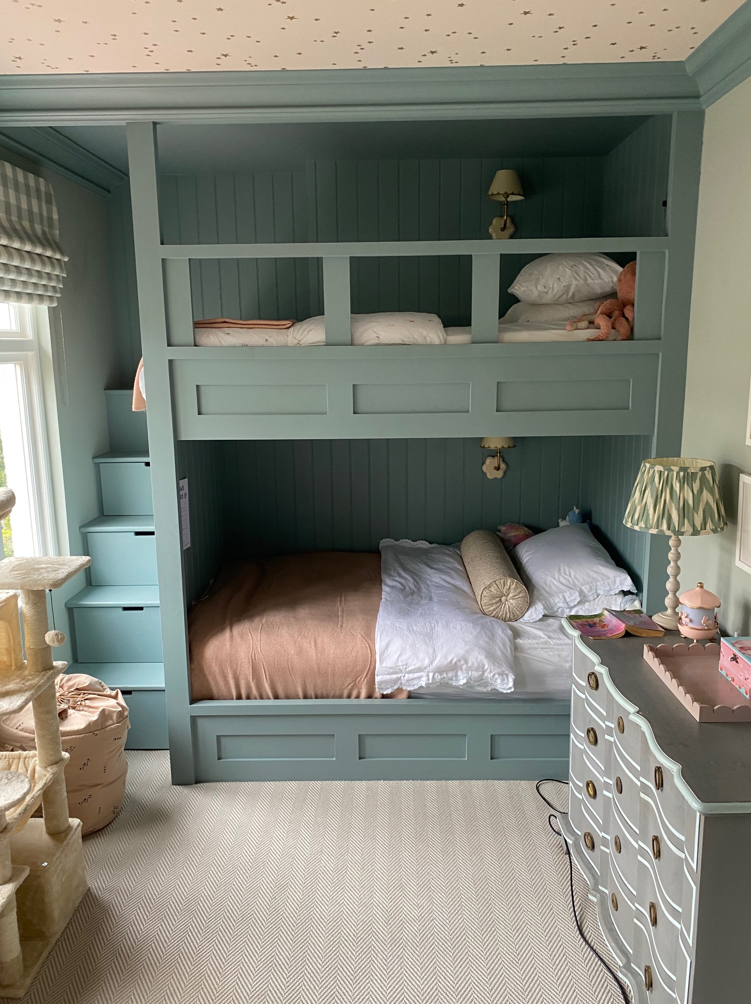 Turkusowe łóżko piętrowe z wbudowanymi schodami-szufladami w pokoju dziecięcym, z wzorzystą tapetą na suficie, obok komoda z falistymi szufladami i lampką nocną.