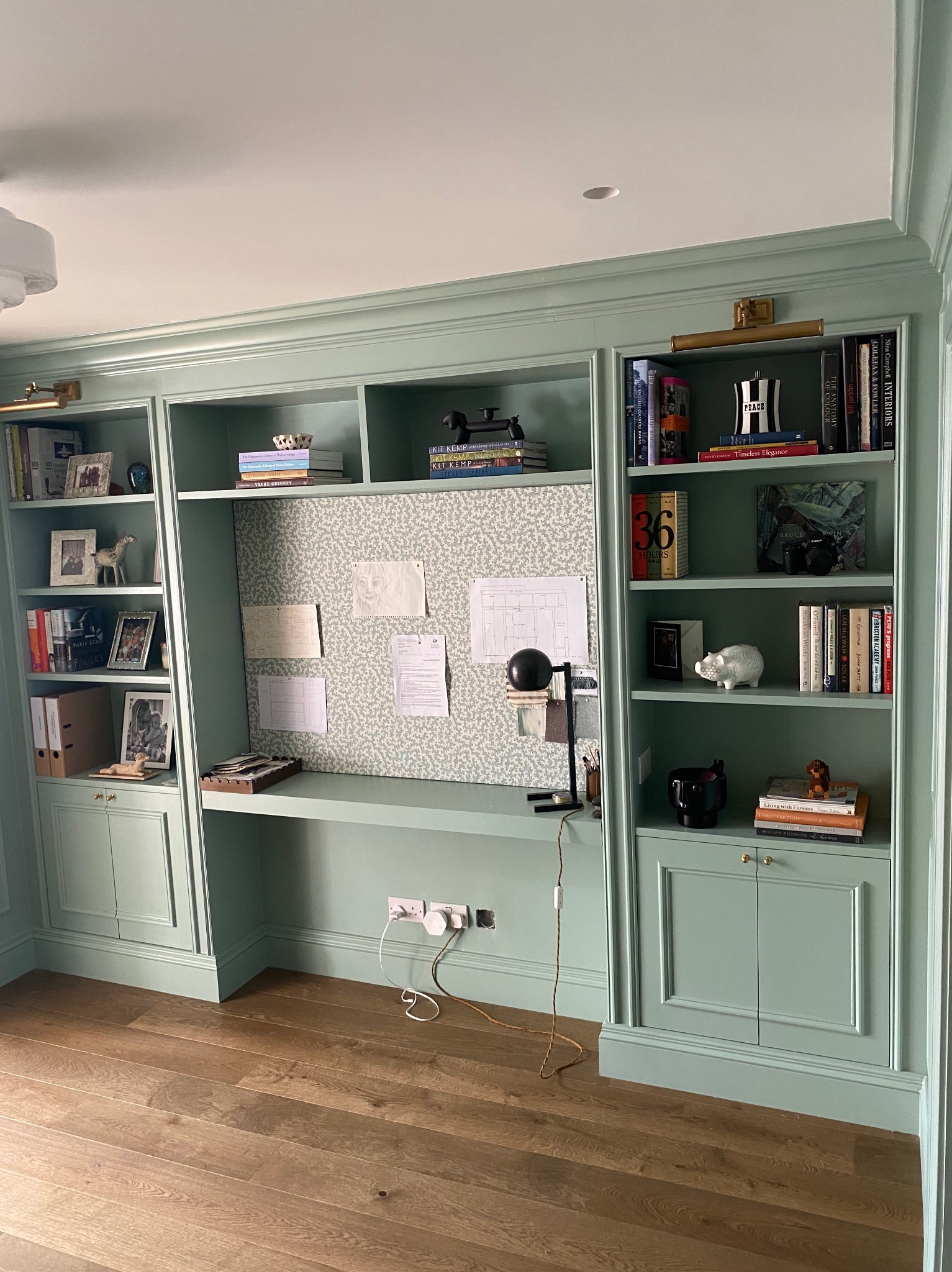 Zabudowa biblioteczna z biurkiem w kolorze miętowym, z półkami wypełnionymi książkami, ramkami ze zdjęciami i dekoracjami, na ścianie tablica korkowa z notatkami, drewniana podłoga.