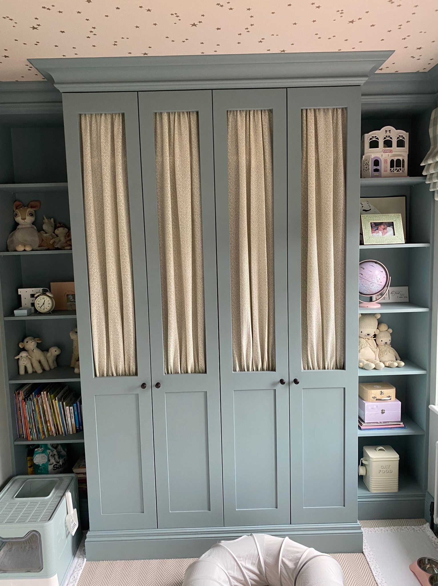 Niebieska zabudowa meblowa z czterema drzwiami z beżowymi zasłonkami, otwartymi półkami z dekoracjami i zabawkami w pokoju dziecięcym.