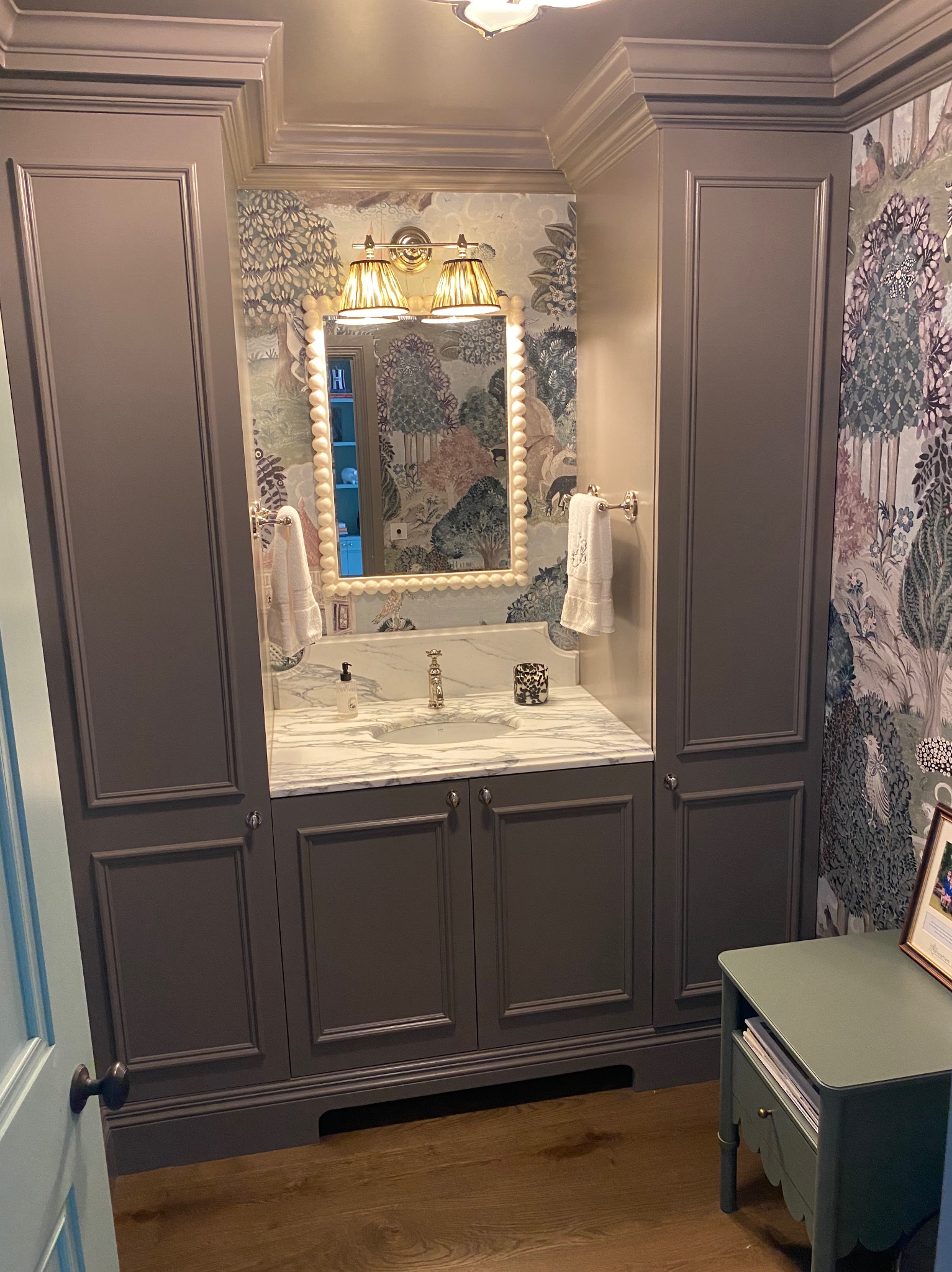 Elegancka łazienka z zabudową szafkową w kolorze taupe, marmurowym blatem umywalkowym, lustrem w ramie z pereł i tapetą w motywy leśne, oświetlona lampami z plisowanymi abażurami.