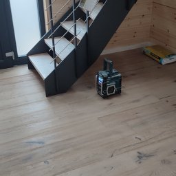 Usługi remontowo budowlane - Jasne panele podłogowe z widoczną strukturą drewna, fragment schodów z metalową konstrukcją i radio budowlane Makita. W tle ściana z drewnianych paneli.