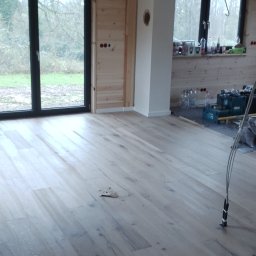 Usługi remontowo budowlane - Jasne panele podłogowe w nowym domu z dużym oknem i drewnianą boazerią. Widoczne narzędzia i materiały budowlane. Podłoga po ułożeniu, gotowa do użytku.