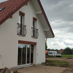 Usługi remontowo budowlane - Boczny widok domu z białą elewacją, czerwonym dachem i ceglanymi detalami wokół okien. Widoczne balustrady balkonowe i przesuwne drzwi tarasowe. W tle krajobraz z przyczepą kempingową.