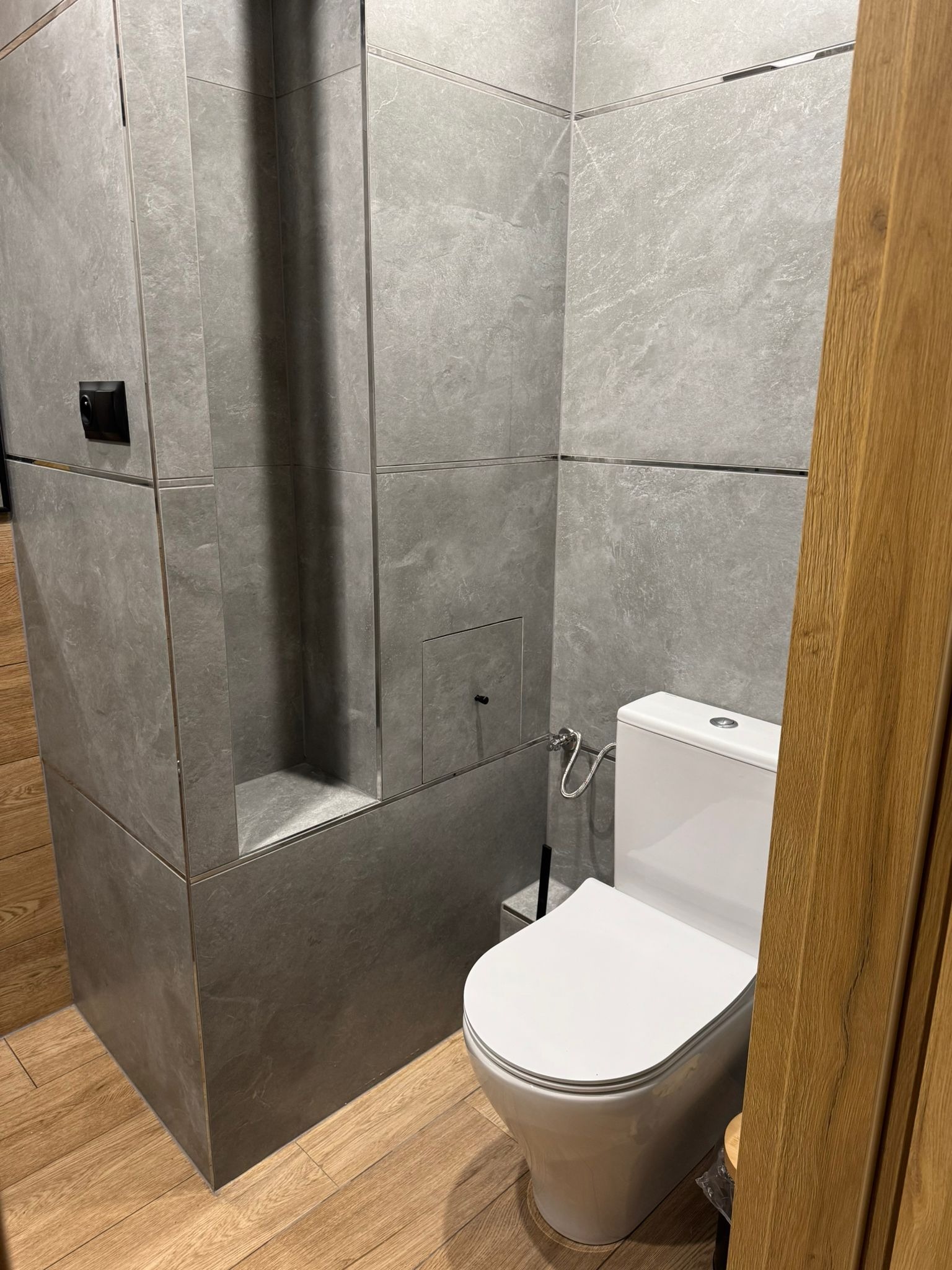 Nowoczesna łazienka z szarymi płytkami imitującymi kamień, drewnianą podłogą i wiszącą toaletą. W ścianie wnęka z półką i ukryty schowek.