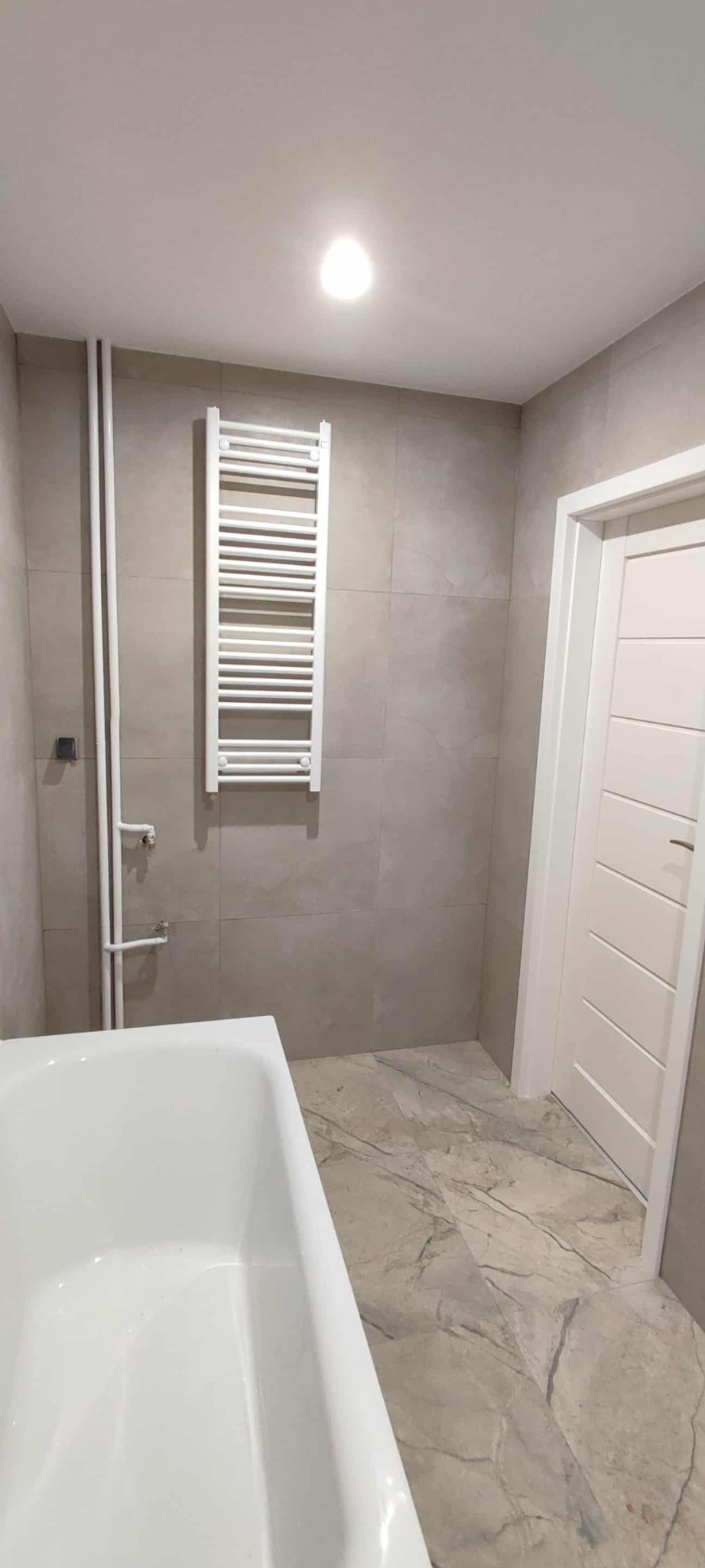 Wykończona łazienka w beżowych płytkach z marmurowym wzorem na podłodze. Widoczna wanna, grzejnik drabinkowy i białe rury. Nowoczesny design.