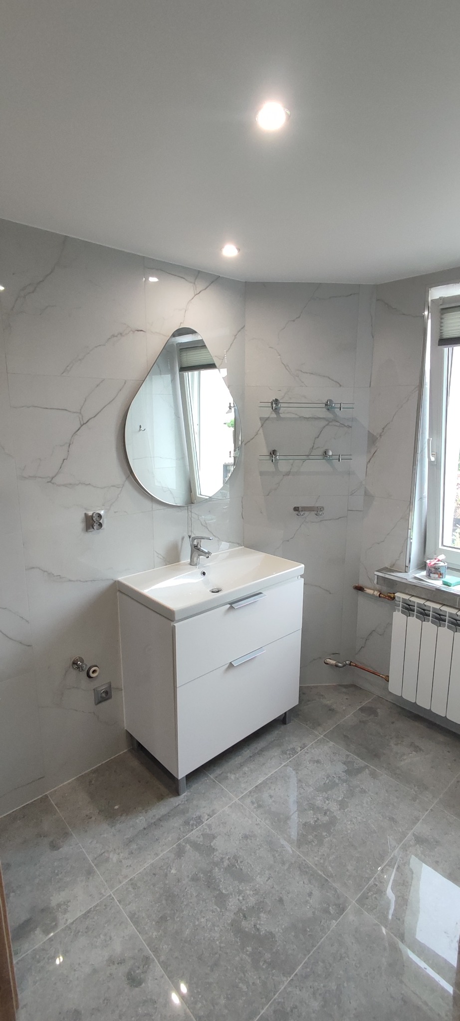 Wykończona łazienka w szaro-białej tonacji: marmurowe płytki ścienne, szara podłoga, biała szafka z umywalką i lustro w kształcie łzy. Nowoczesny design.