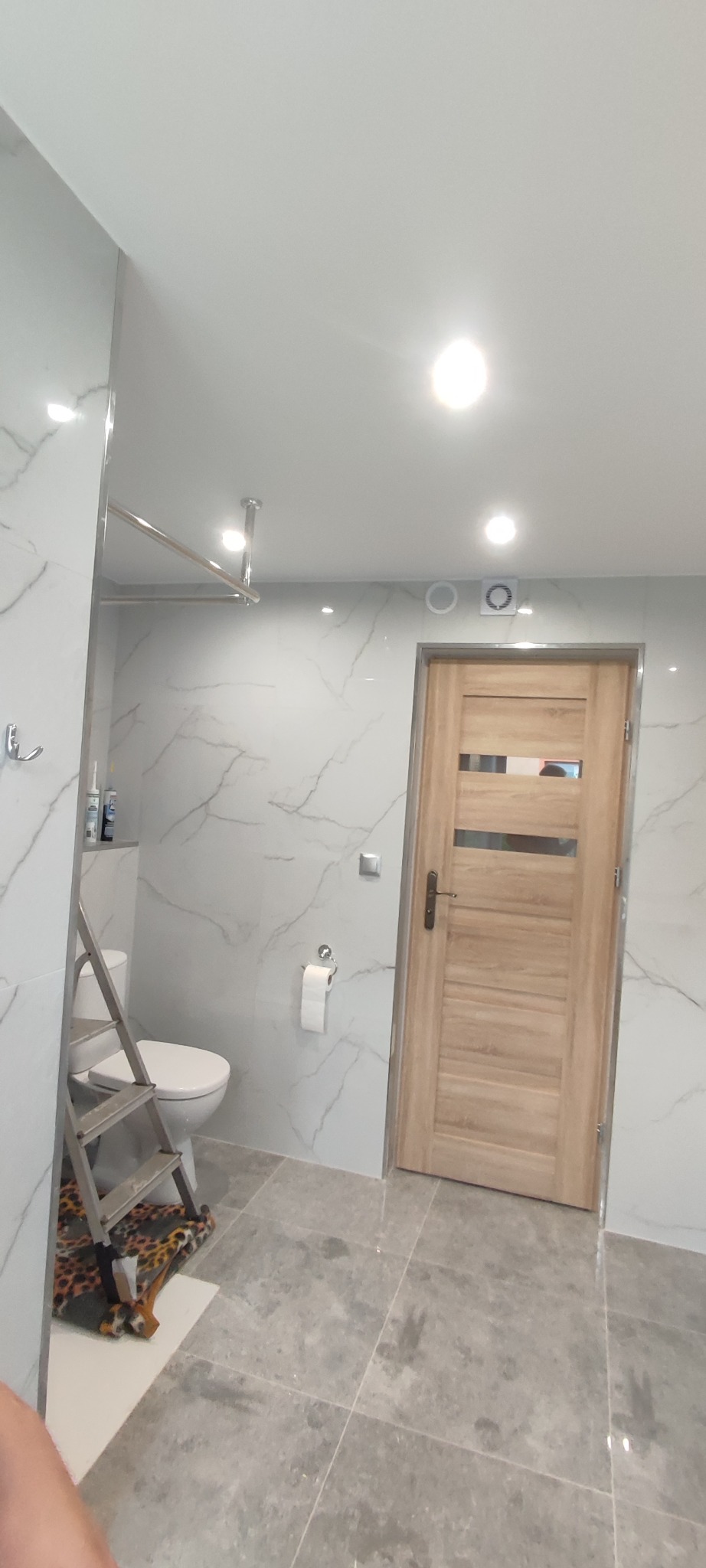 Wykończona łazienka z szarymi płytkami imitującymi marmur na ścianach i podłodze. Widoczna drabina oparta o toaletę, sugerująca trwające prace wykończeniowe.