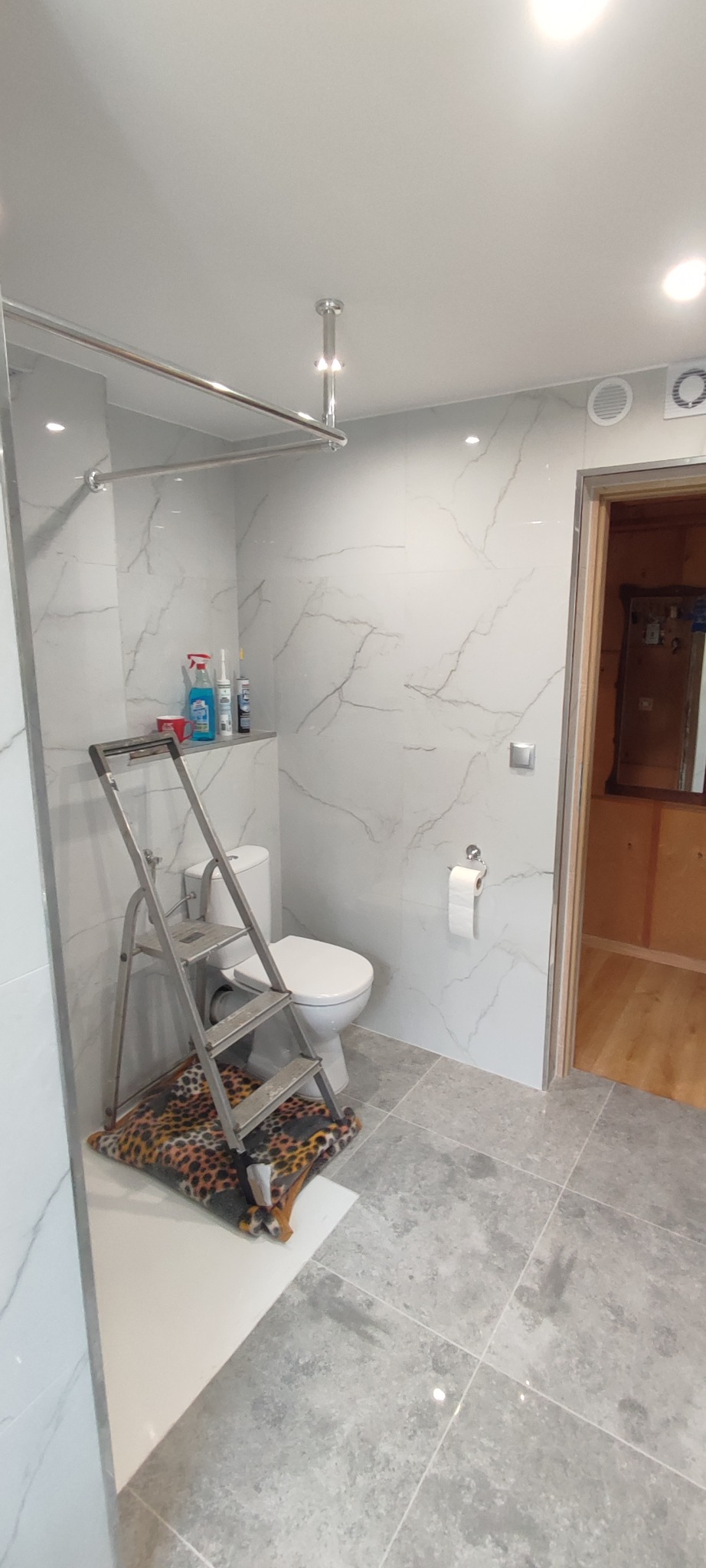 Wykończona łazienka z marmurowymi płytkami, widoczna drabina malarska, toaleta i akcesoria. W tle drewniane drzwi do innego pomieszczenia. Nowoczesny design.