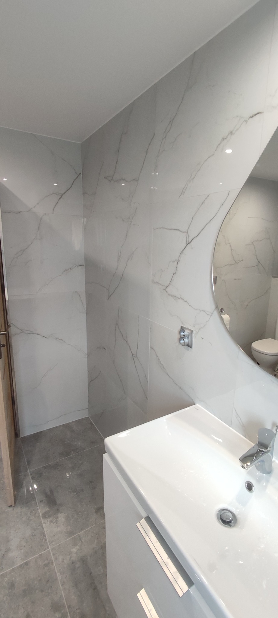 Nowoczesna łazienka po remoncie: białe marmurowe płytki na ścianach, szara podłoga, okrągłe lustro i biała umywalka z szafką. Widoczny fragment toalety w odbiciu lustra.