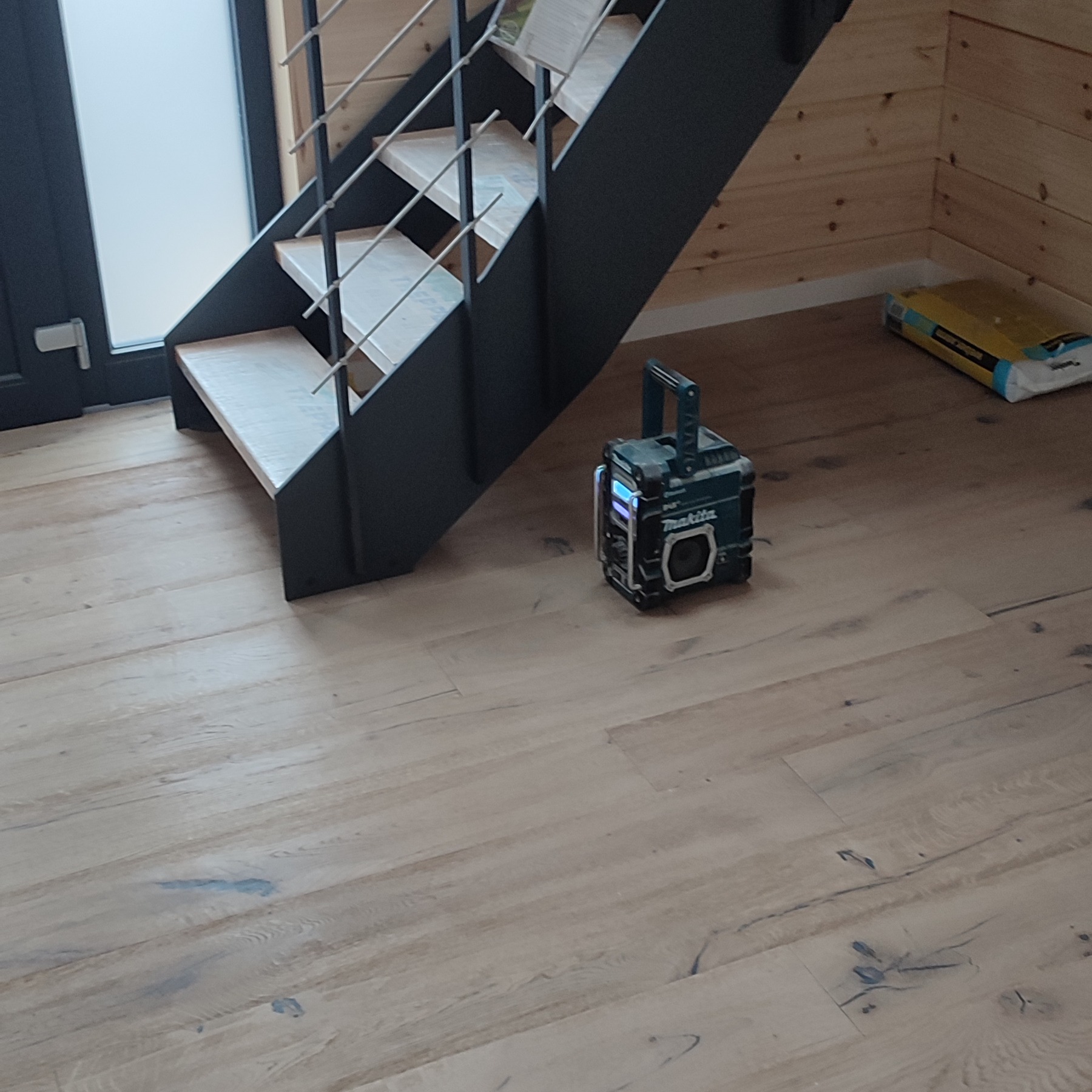 Jasne panele podłogowe z widoczną strukturą drewna, fragment schodów z metalową konstrukcją i radio budowlane Makita. W tle ściana z drewnianych paneli.