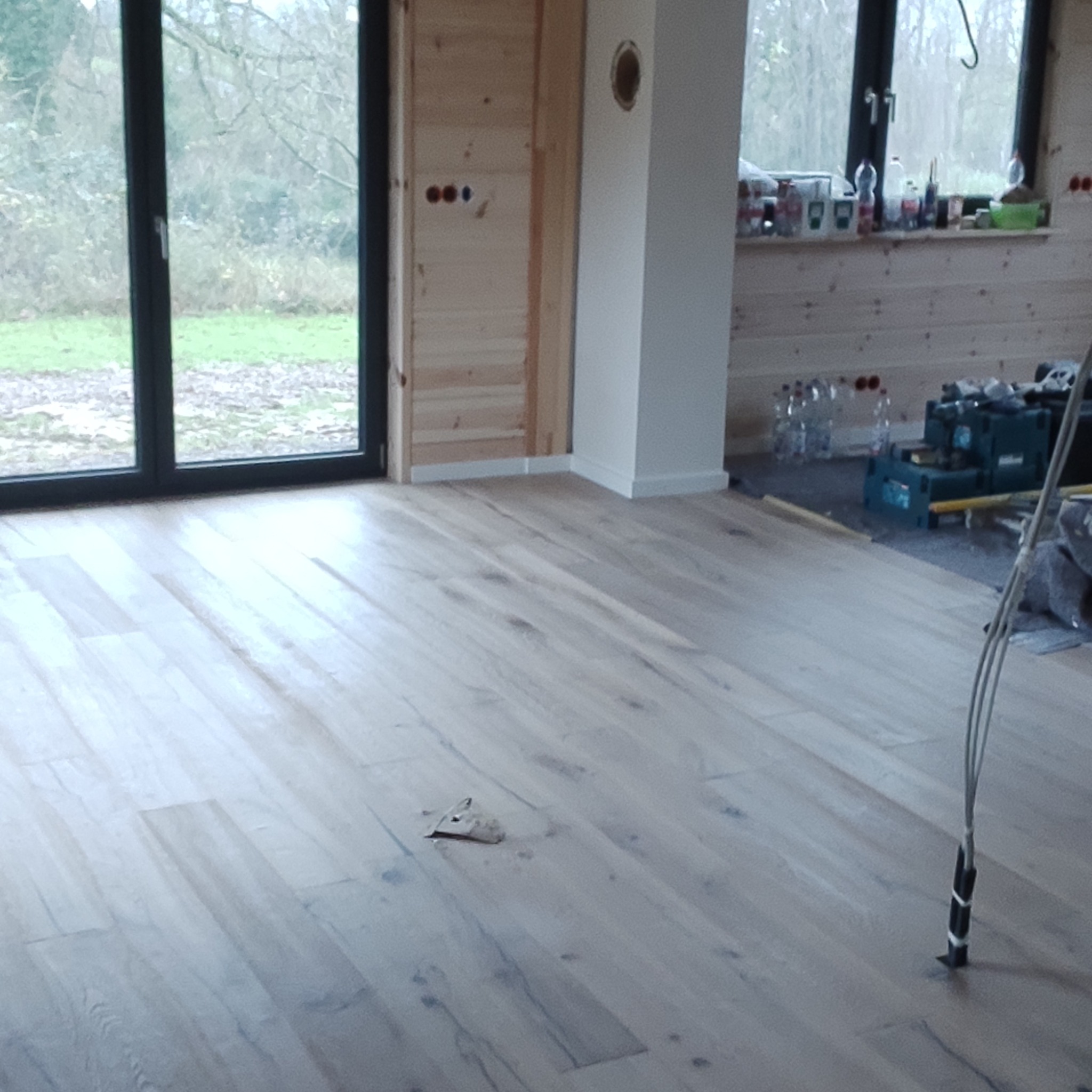 Jasne panele podłogowe w nowym domu z dużym oknem i drewnianą boazerią. Widoczne narzędzia i materiały budowlane. Podłoga po ułożeniu, gotowa do użytku.