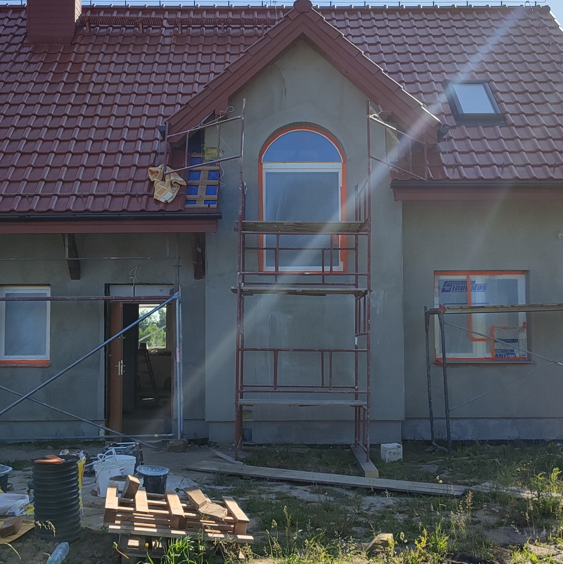 Dom jednorodzinny w trakcie prac elewacyjnych: szare ściany, rusztowania, zabezpieczone okna folią, czerwona dachówka. Widoczne materiały budowlane.
