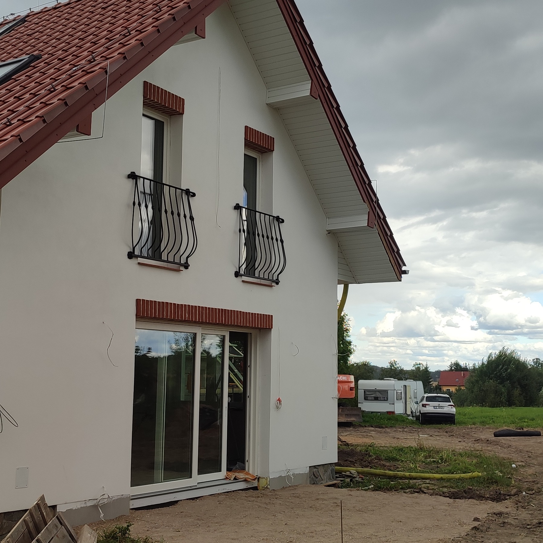 Boczny widok domu z białą elewacją, czerwonym dachem i ceglanymi detalami wokół okien. Widoczne balustrady balkonowe i przesuwne drzwi tarasowe. W tle krajobraz z przyczepą kempingową.